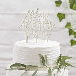 Wooden cake topper featuring the phrase "And So The Adventure Begins" in elegant script, measuring 22cm x 15cm, ideal for wedding or engagement cakes.